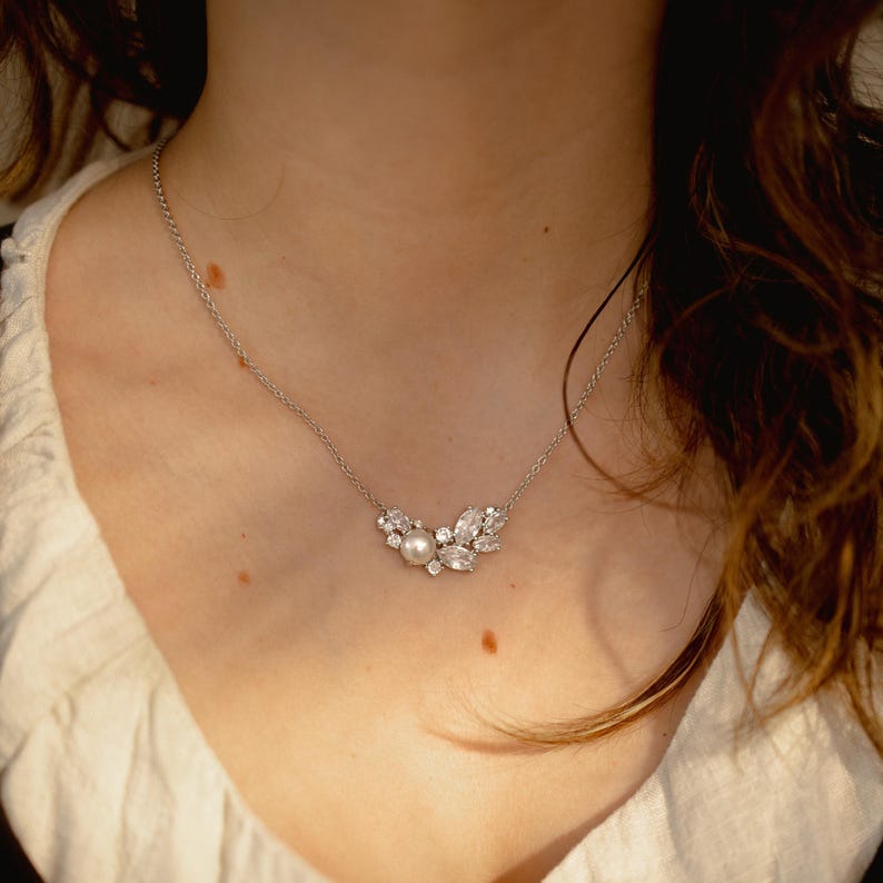 May include: A silver necklace with a cluster pendant. The pendant features a central pearl surrounded by clear, sparkling crystals. The necklace is delicate and sits on a person's chest.
