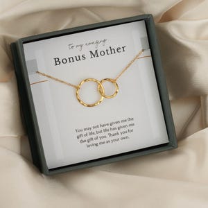 Pode incluir: Um colar dourado com dois círculos interligados, apresentado numa caixa cinza escuro. A caixa contém um cartão com o texto "To my amazing Bonus Mother" e uma mensagem comovente. O colar repousa sobre um tecido creme.