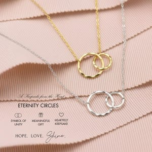 A necklace with two intertwined rings, one gold and one silver, against a pink fabric background.
