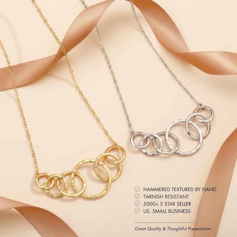 May include: Two necklaces with linked circles, one gold-toned and one silver-toned, are displayed against a neutral background with a ribbon. The gold necklace is on the left, and the silver necklace is on the right. The necklaces have a hammered texture.