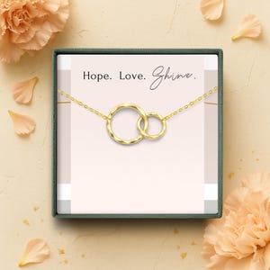 May include: A gold-colored necklace with two interlocking circles, presented in a gift box. The box features the words "Hope. Love. Shine." The necklace is displayed against a light pink background, with floral accents.