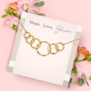 May include: A gold-colored necklace with six interconnected circles, displayed on a card with the words "Hope. Love. Shine." The necklace is set against a pink background with floral accents, creating a delicate and elegant aesthetic.