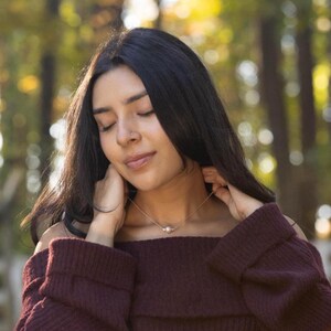 May include: A gold-colored necklace with a single pearl pendant. The necklace is worn on a person with dark hair and a burgundy sweater. The background is a blurred view of a forest with sunlight.