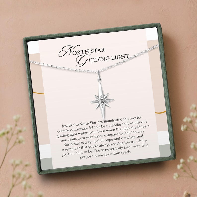 K&ouml;nnte beinhalten: Eine silberne Halskette mit einem Nordstern-Anh&auml;nger, pr&auml;sentiert in einer Geschenkbox. Der Anh&auml;nger hat ein Sternenmuster mit einem zentralen Edelstein. Der Text "North Star Guiding Light" ist auf der Karte aufgedruckt.