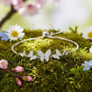 May include: A silver butterfly bracelet with sparkling crystals rests on a bed of green moss, surrounded by daisies, pink cherry blossoms, and blue forget-me-nots. The bracelet features three butterfly charms.