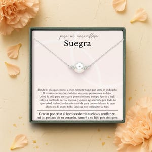 Peut inclure: Un collier en argent avec un grand pendentif perle blanche et de petits accents décoratifs. Le collier est présenté dans un coffret cadeau avec du texte en espagnol, dont le mot "Suegra". Le fond présente des accents floraux.
