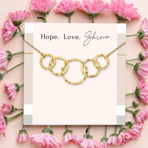 Può includere: Una collana dorata con cinque cerchi interconnessi, su uno sfondo rosa tenue con fiori rosa. Le parole "Hope. Love. Shine." sono stampate sopra la collana.