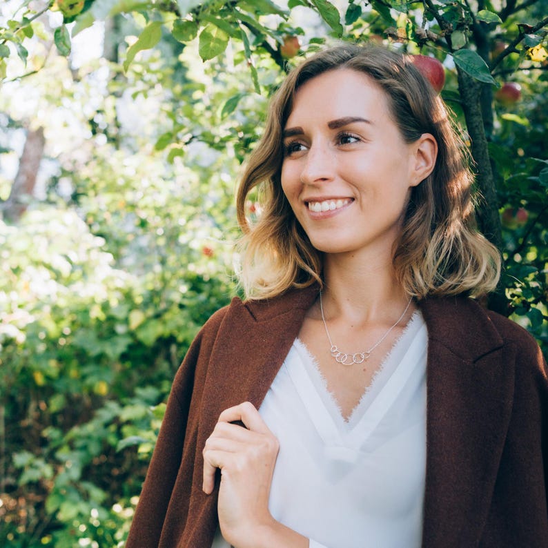 May include: A woman wearing a brown coat and a white blouse with a lace trim. A silver necklace with a circular design is visible. The background features green foliage and sunlight.