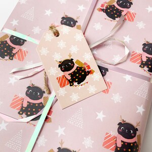 May include: Pink wrapping paper with a repeating pattern of a black pug wearing a scarf and antlers, surrounded by gifts, snowflakes, and Christmas trees. A matching gift tag is attached with twine and iridescent ribbon.