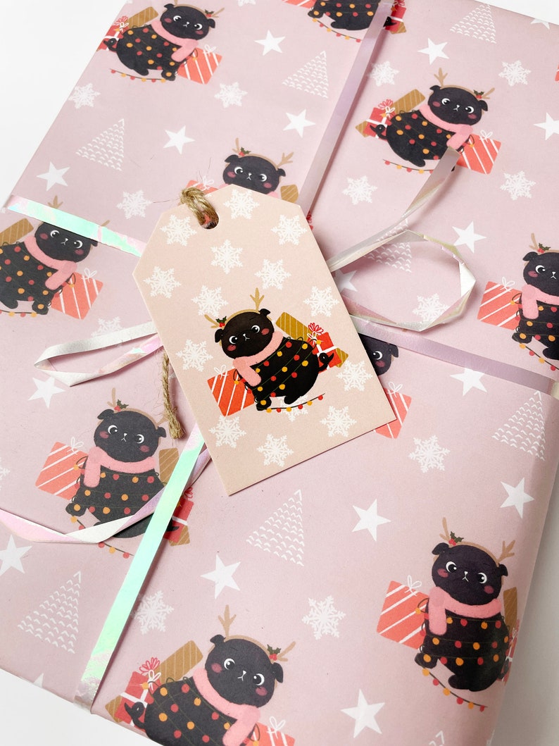 May include: Pink wrapping paper with a repeating pattern of a black pug wearing a scarf and antlers, surrounded by gifts, snowflakes, and Christmas trees. A matching gift tag is attached with twine and iridescent ribbon.
