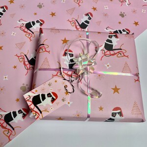 May include: A wrapped gift with pink wrapping paper featuring a repeating pattern of black and white cats wearing Santa hats, gold stars, and Christmas trees. The gift is tied with a silver ribbon and a holographic bow, and includes a matching gift tag.