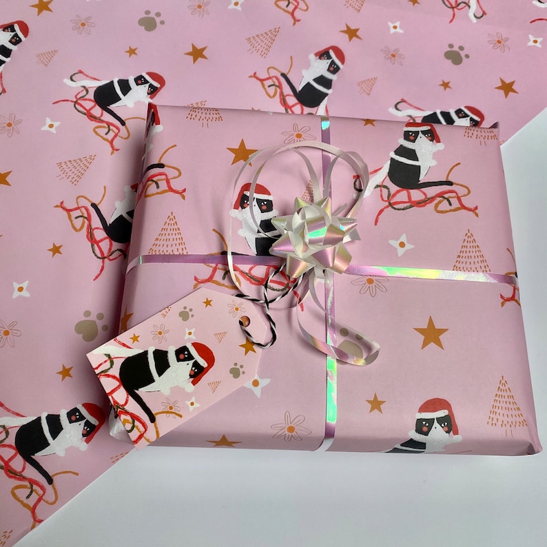 May include: A wrapped gift with pink wrapping paper featuring a repeating pattern of black and white cats wearing Santa hats, gold stars, and Christmas trees. The gift is tied with a silver ribbon and a holographic bow, and includes a matching gift tag.