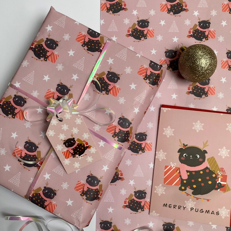 May include: Pink wrapping paper, gift tag, and greeting card featuring a repeating pattern of pugs wearing scarves and holding presents. The card reads "MERRY PUGMAS". A gold glitter ornament and ribbon are also present.