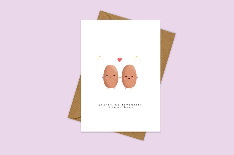 May include: A white card with two brown cartoon beans holding hands. The beans have happy faces and there is a red heart above them. The text on the card reads "You're my favourite human bean."
