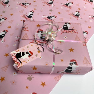 May include: A wrapped gift with pink wrapping paper featuring a repeating pattern of a dog wearing a Santa hat on a sled, with gold stars, paw prints, and flowers. The gift is tied with a silver bow and a matching gift tag.