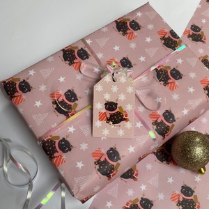 May include: Pink wrapping paper with a repeating pattern of a black pug wearing a scarf and reindeer antlers, surrounded by gifts, stars, and Christmas trees. A matching gift tag is attached with a ribbon.