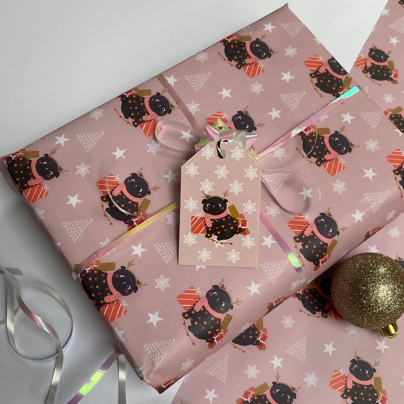 May include: Pink wrapping paper with a repeating pattern of a black pug wearing a scarf and reindeer antlers, surrounded by gifts, stars, and Christmas trees. A matching gift tag is attached with a ribbon.