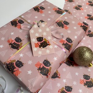May include: Pink wrapping paper with a repeating pattern of a black pug wearing a pink scarf and reindeer antlers, holding a gift. White stars and Christmas trees are also on the paper. A matching gift tag is attached with a silver ribbon.