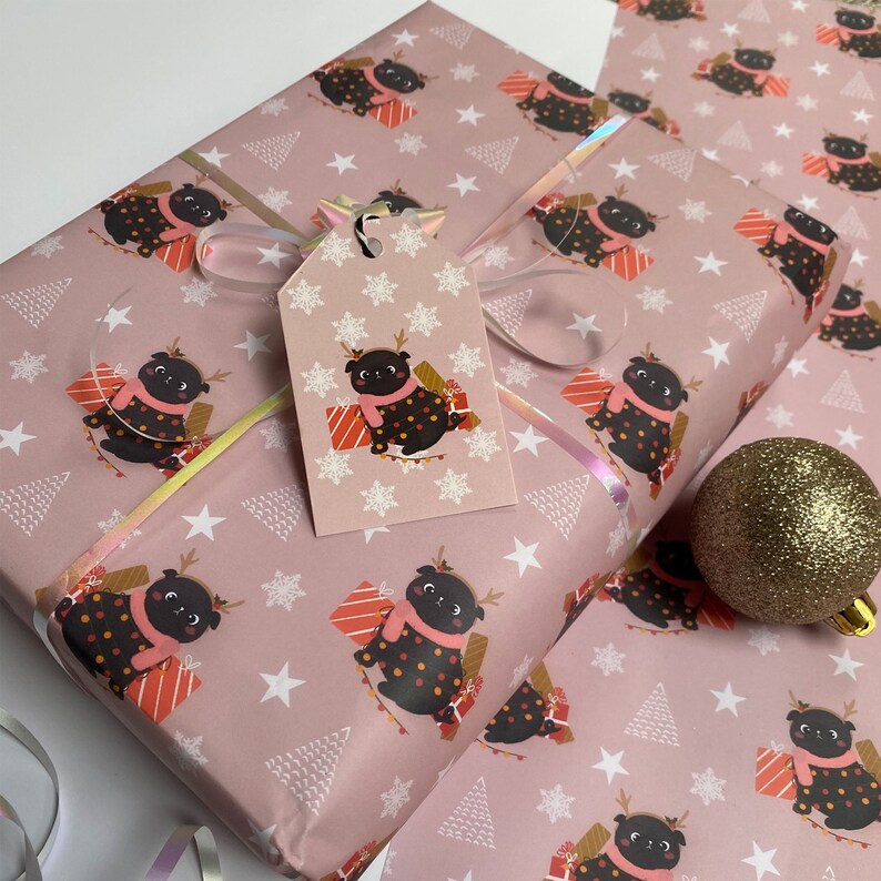 May include: Pink wrapping paper with a repeating pattern of a black pug wearing a pink scarf and reindeer antlers, holding a gift. White stars and Christmas trees are also on the paper. A matching gift tag is attached with a silver ribbon.