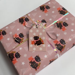 May include: A wrapped gift with pink wrapping paper featuring a repeating pattern of a pug wearing a scarf and reindeer antlers, holding a present. A matching gift tag is attached with a holographic ribbon and bow.