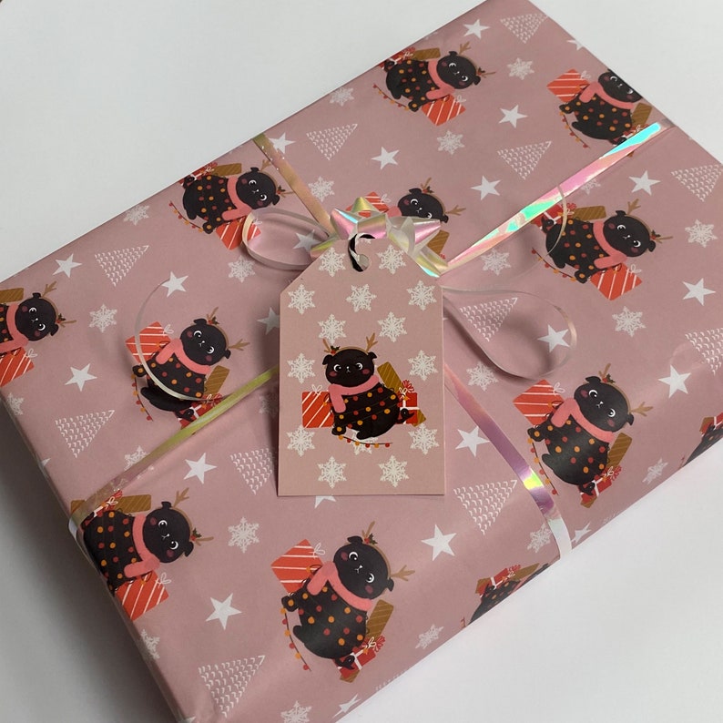 May include: A wrapped gift with pink wrapping paper featuring a repeating pattern of a pug wearing a scarf and reindeer antlers, holding a present. A matching gift tag is attached with a holographic ribbon and bow.