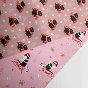 May include: Two sheets of wrapping paper with festive patterns. One sheet features a repeating pattern of black pugs wearing reindeer antlers and holding gifts on a dusty rose background. The other sheet has a pink background with a cat wearing a Santa hat.