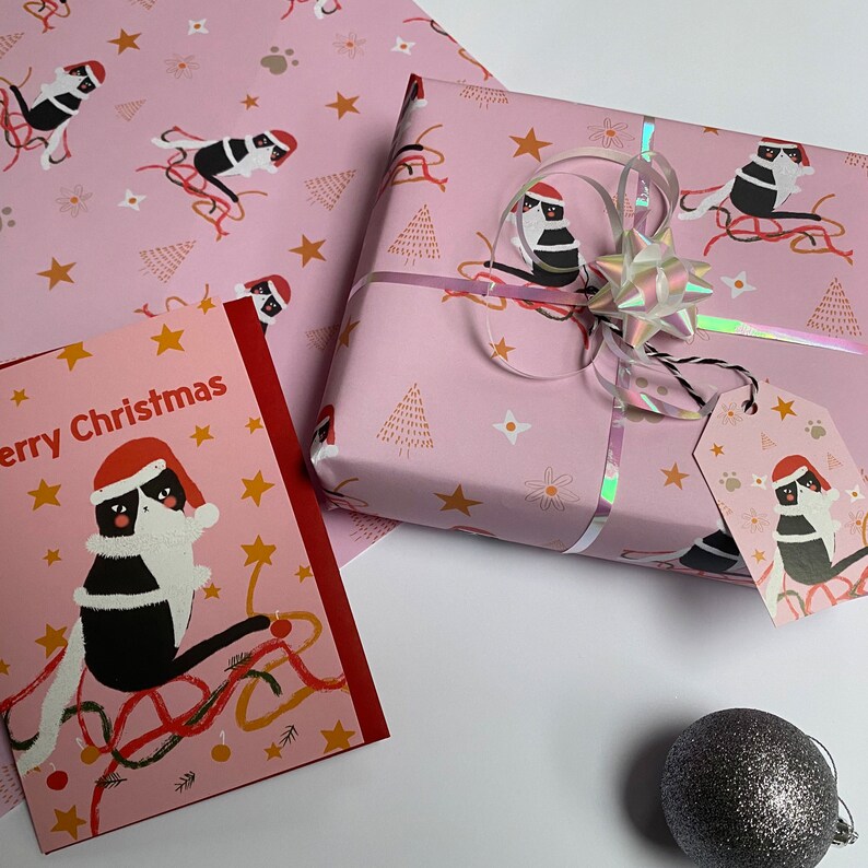 May include: Pink Christmas-themed wrapping paper, gift tag, and card featuring a black and white cat wearing a Santa hat. The wrapping paper has a repeating pattern of cats, stars, and Christmas trees. A wrapped gift is tied with a holographic ribbon and bow.