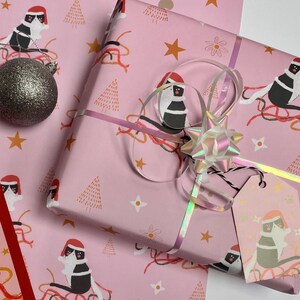May include: Pink wrapping paper with a repeating pattern of cats wearing Santa hats, Christmas trees, stars, and flowers. A wrapped gift is tied with a holographic ribbon and bow, and a gift tag is attached. A silver glitter ornament is next to the gift.