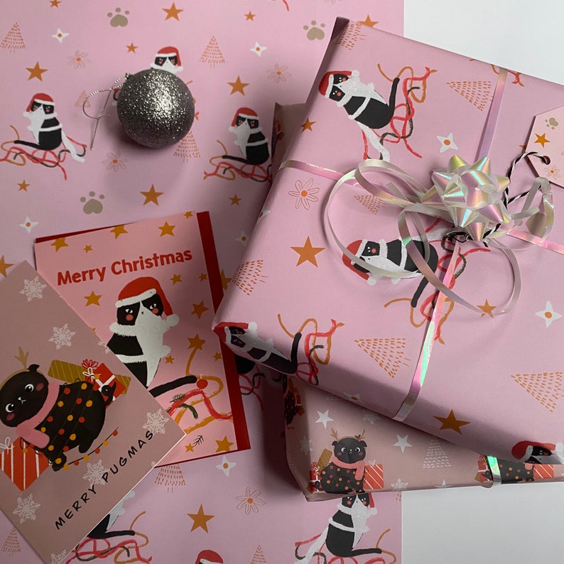 May include: Pink Christmas-themed wrapping paper, gift boxes, and greeting cards. The wrapping paper and cards feature a repeating pattern of pugs wearing Santa hats and riding sleds. The cards read "Merry Christmas" and "Merry Pugmas". A silver glitter ornament and a holographic bow are also present.