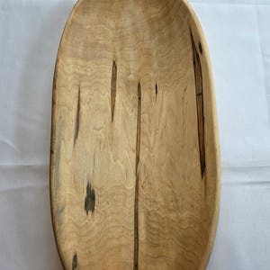 May include: Oval wooden bowl with a natural finish. The bowl has a light brown color with dark streaks and knots, showcasing the wood grain. The elongated shape is ideal for serving or display.