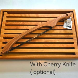 Puede incluir: Una tabla de pan de madera con un cuchillo de cerezo. La tabla es rectangular con ranuras paralelas y un borde elevado. El cuchillo tiene un mango de madera curvado y una hoja negra. El texto "With Cherry Knife (optional)" es visible.