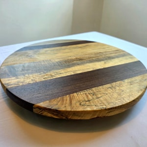 May include: A circular wooden serving board with alternating light and dark wood grain stripes. The board is approximately 30 cm in diameter, featuring a dark brown band of wood in the centre. The edges are rounded and finished, displaying the natural wood patterns.