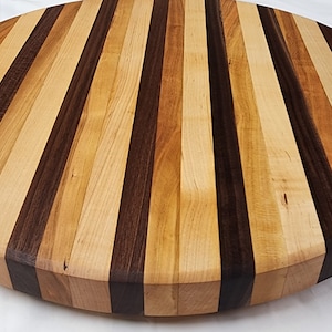 Puede incluir: Una tabla de cortar redonda de madera con un diseño a rayas. La tabla está hecha de diferentes tipos de madera, incluyendo arce, nogal y cerezo. La tabla tiene un diámetro de aproximadamente 30 cm.