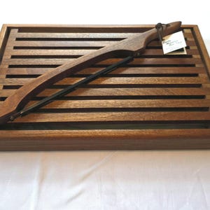 May include: A rectangular wooden bread slicing board with a slatted surface and a matching wooden bread knife featuring a black metal blade. The knife handle is attached to the board with a small leather cord and a white paper product tag.