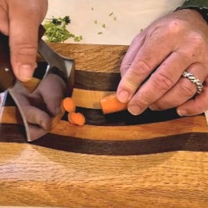 Peut inclure: Une personne utilisant un hachoir &agrave; l&eacute;gumes en acier inoxydable pour hacher des carottes sur une planche &agrave; d&eacute;couper en bois avec un design incurv&eacute;. La planche &agrave; d&eacute;couper a un centre en bois fonc&eacute; avec des bandes de bois plus claires de chaque c&ocirc;t&eacute;.