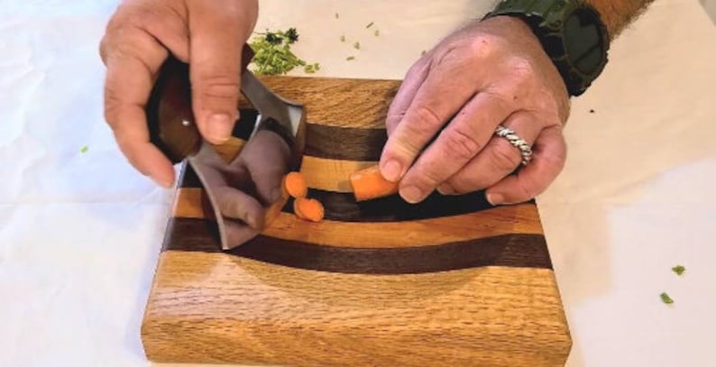 Peut inclure: Une personne utilisant un hachoir &agrave; l&eacute;gumes en acier inoxydable pour hacher des carottes sur une planche &agrave; d&eacute;couper en bois avec un design incurv&eacute;. La planche &agrave; d&eacute;couper a un centre en bois fonc&eacute; avec des bandes de bois plus claires de chaque c&ocirc;t&eacute;.