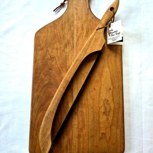 Puede incluir: Una tabla de cortar de madera con un cuchillo de arco de cerezo a juego. La tabla tiene un asa y un lazo de cuero para colgar. El cuchillo tiene una hoja curva y un mango de madera. El texto "with Optional Cherry Bow Knife" está en la parte inferior.