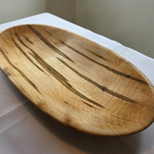 May include: Oval wooden bowl with a natural wood grain pattern. The bowl is light brown with dark brown streaks running lengthwise. The bowl is sitting on a white surface.