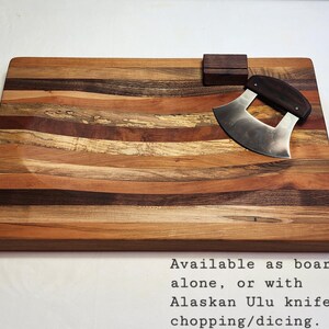 Peut inclure: Une planche &agrave; d&eacute;couper en bois avec une surface incurv&eacute;e et rainur&eacute;e. La planche est faite de plusieurs types de bois, cr&eacute;ant un motif ray&eacute;. Un petit couteau incurv&eacute; avec un manche en bois est plac&eacute; sur la planche. Le texte "Available as board alone, or with Alaskan Ulu knife for chopping/dicing." est imprim&eacute; sous la planche.