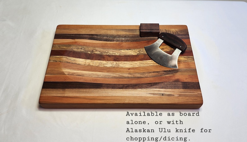 Peut inclure: Une planche &agrave; d&eacute;couper en bois avec une surface incurv&eacute;e et rainur&eacute;e. La planche est faite de plusieurs types de bois, cr&eacute;ant un motif ray&eacute;. Un petit couteau incurv&eacute; avec un manche en bois est plac&eacute; sur la planche. Le texte "Available as board alone, or with Alaskan Ulu knife for chopping/dicing." est imprim&eacute; sous la planche.