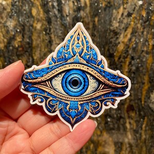 May include: A blue and gold eye sticker with intricate details. The eye is surrounded by a decorative mandala-like design.