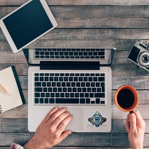 May include: A silver laptop computer with a blue and gold eye sticker on the trackpad. The laptop is on a wooden table with a notebook, pencil, coffee cup, and camera.