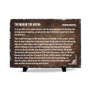 May include: A rectangular slate plaque with a dark wood grain background featuring the white text of Theodore Roosevelt's The Man in the Arena speech. The plaque has rough, natural stone edges and is supported by two small black plastic stands.