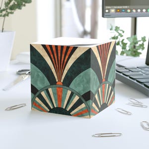 May include: A cube-shaped notepad holder with an Art Deco design. The pattern features teal, orange, black, and beige fan-like shapes. The notepad holder sits on a white surface with scattered silver paper clips.