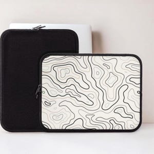 May include: A rectangular laptop sleeve featuring a white background with black and light gray topographic map contour lines. The sleeve has a black zipper closure and black trim along the edges. A second solid black laptop sleeve is visible behind it.