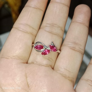 May include: A silver ring with three marquise-cut, vivid red gemstones. The ring's design incorporates a looping detail and a stylized leaf or petal arrangement, creating an elegant and striking piece of jewellery.