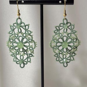 May include: A pair of sage green tatting lace earrings featuring intricate knotted thread patterns, small light green seed beads, and round light green gemstone accents. The earrings are suspended from gold-toned metal fishhook ear wires.