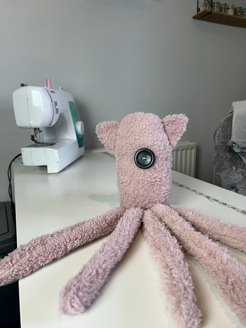 May include: A plush pink octopus toy with a large black button eye. The stuffed animal has eight long tentacles and small ear-like appendages on its head. A white sewing machine is in the background.