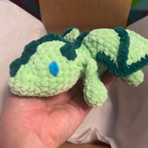 Puede incluir: Un peque&ntilde;o drag&oacute;n de peluche tejido a ganchillo a mano con hilo suave de color verde menta claro. Presenta detalles en verde bosque oscuro a lo largo de la columna y las alas, con ojos ovalados de color azul brillante cosidos a los lados.