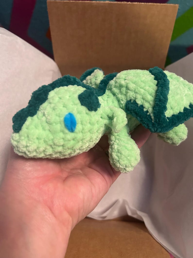 Puede incluir: Un peque&ntilde;o drag&oacute;n de peluche tejido a ganchillo a mano con hilo suave de color verde menta claro. Presenta detalles en verde bosque oscuro a lo largo de la columna y las alas, con ojos ovalados de color azul brillante cosidos a los lados.
